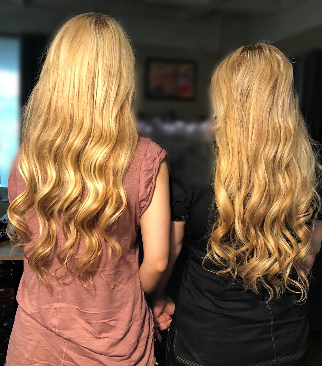 Bakifrånvy av två individer med långt, vågigt blont hår. Personen till vänster bär en rosa/rosé topp medan personen till höger bär en svart topp. Båda har liknande gyllenblont hår med strandvågor som når mitten av ryggen.